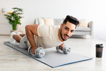 Stay at home fitness. Strong young Arab man doing whole body workout, standing in plank pose, using dumbbells indoors