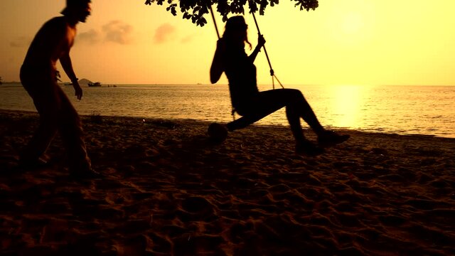 Man is pushing woman sitting on a swing to sway her on a beach with sunset and ocean on background. Couple relaxes near water at dusk enjoying warm weather and tropical nature. Vacations on a seashore