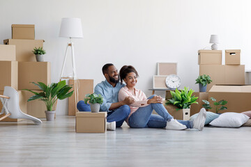 Cheerful young black husband and wife rest, sit on floor among cardboard boxes with things and plants