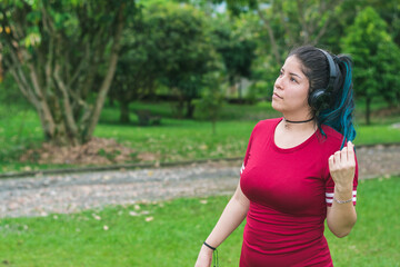 beautiful college woman with blue hair and red dress, listening to music with her wireless headphones in a park, looking slightly upward as she touches her hair. woman analyzing options.