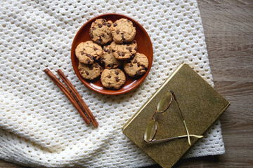 Plate of chocolate chip cookies, knitted blanket, book, reading glasses and cinnamon sticks. Hygge at home. Flat lay.