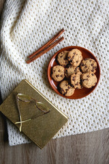 Plate of chocolate chip cookies, knitted blanket, book, reading glasses and cinnamon sticks. Hygge at home. Flat lay.