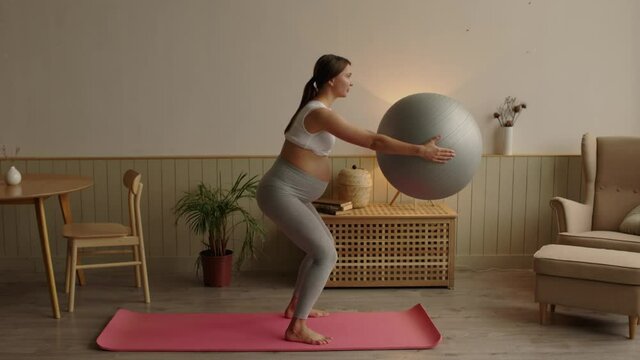Pregnant woman exercising with fit ball at home, making squats with fitball.
