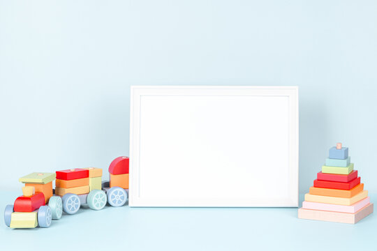 Toys background. White wooden picture frame with blank mock up copy space standing next to wooden train and colorful toy pyramid on pastel blue background. Front view