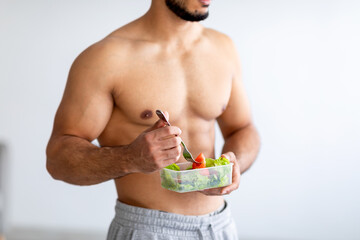 Balanced diet concept. Cropped view of strong young man with bare torso eating vegetable salad at...