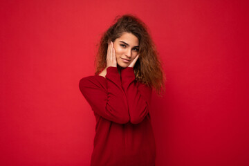 Young beautiful self-confident curly brunette woman with sincere emotions poising isolated on background wall with copy space wearing casual dark red sweater. Positive concept