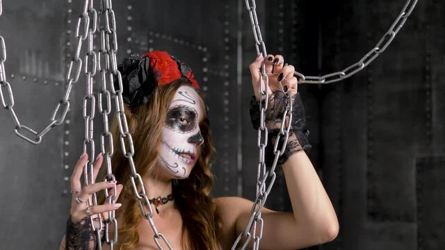 Lady model with Halloween makeup in wreath choker and in lacy gloves poses with chains hanging from ceiling against studio walls closeup