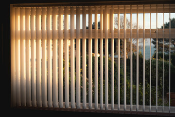 Vertical blinds with a golden glow as the sun sets.There is a tree and a view of the sea through the window.