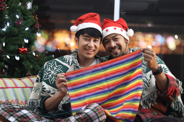 Adorable smiling young LGBT couple share blanket, hold rainbow flag, two men have special moment together on Christmas holiday, Asian gay lover sit on sofa in living room to celebrate Christmas night