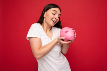 Adorable positive smiling young beautiful charming brunette woman with sincere emotions wearing casual white t-shirt isolated over red background with copy space and holding pink piggy box. Moneybox