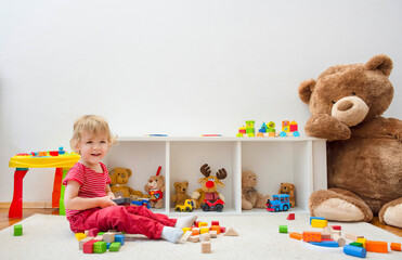 Sweet happy child boy having fun at home playing with his giant teddy bear and colorful wooden blocks and toys, on the floor.