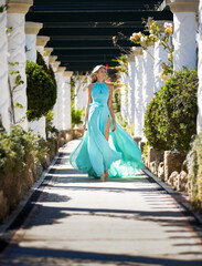 Beautiful blonde milf woman comes in long turquoise flying dress among white columnns along a...
