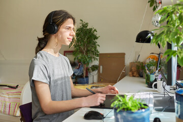 Teenage guy in headphones sitting at home using computer and a graphics tablet