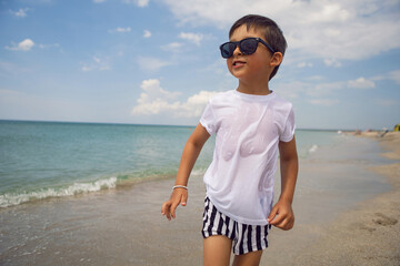 boy child in striped shorts and a white T-shirt walks on sandy beach and in sunglasses