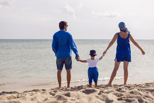 Family Of Three In Blue Clothes Stands Back On A Sandy Beach The Sea In Summer On Vacation