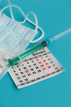 a calendar sheet with the circled date of November 17 on a blurry background with a disposable syringe, a medical mask and white tablets on a blue background in vertical format