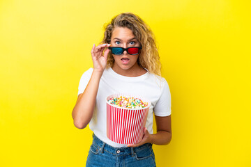Obraz premium Girl with curly hair isolated on yellow background surprised with 3d glasses and holding a big bucket of popcorns