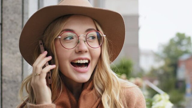 Portrait attractive young girl talking on phone outdoors close-up caucasian happy woman uses smartphone to communicate millennial female with hat and glasses smiling holding gadget makes online order