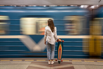 Young girl passenger with longboard standing on subway station platform with blurry moving blue...