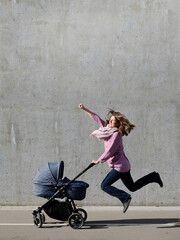 Happy cheerful super heroine mother flying with newborn stroller on grey concrete wall background....