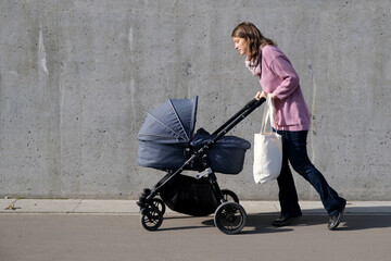 Mother with stroller and canvas eco shopping bag walking near grey concrete wall in the city. Caring mom looking on newborn in baby carriage.