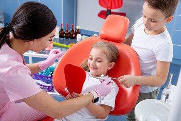 pediatric dentist examines the dental cavity of a little girl. boy helps little girl not to be...