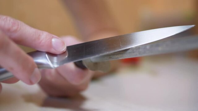 Female Hands Sharpens a Knife on Old, Dirty, Scratched a Steel Rod in Kitchen. Dull knife blade. Preparation of the working tool for cutting. Blurred background. Daylight illumination. 4K. Close up.