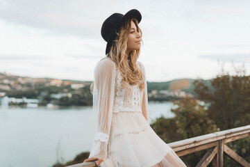 A girl with blond hair in a white boho dress and a hat sits on the observation deck near a turquoise lake