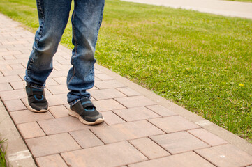 A child in blue jeans and dark sneakers with blue inserts is walking on a sidewalk tile against a background of green grass