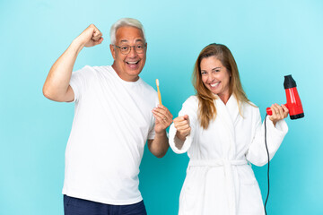 Middle age couple holding dryer and toothbrush isolated on blue background celebrating a victory in winner position