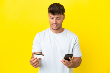 Young caucasian handsome man isolated on yellow background buying with the mobile with a credit card