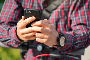 Young man wearing shirt leaning on electric scooter handlebar, holding mobile smartphone in his hands, closeup detail