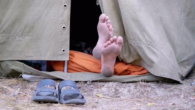 Bare Feet Male Legs Peeking Out from an Open Military Tent in Nature. Tourist is resting, sleeping at camping, airing naked legs. Traveler relaxes in a tent without slippers among trees outdoors. 4K.