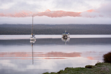 A cold early morning on lake Te Anau, New Zealand with mountains in the background reflected in the still water.