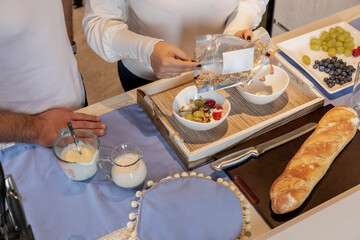 Woman's hands emptying granola into a bowl with yogurt while preparing breakfast with her husband