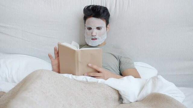 Carefree guy wearing cosmetic face mask and pajamas is reading book in bed at home relaxing enjoying literature and skin care. Youth and lifestyle concept.