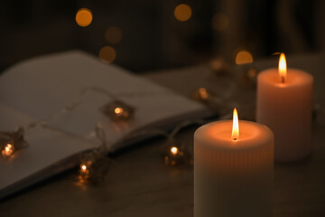 Burning candles, festive lights and open book in darkness, closeup. Space for text