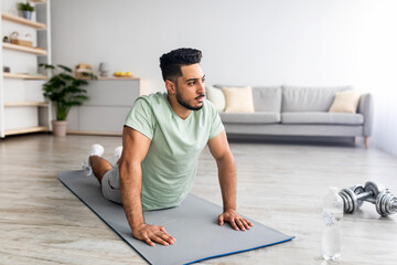 Naklejka na ściany i meble Domestic yoga practice. Flexible young Arab man doing cobra pose on mat, making exercises at home, free space