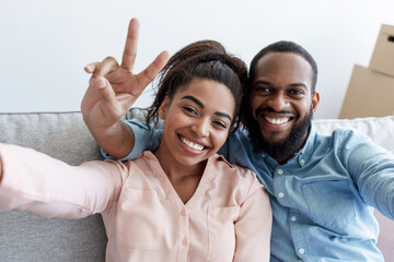 Smiling young black couple rest on couch, man shows peace sign, makes selfie in room