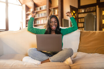 Young black woman with laptop lifting hands up, gesturing YES, celebrating success, winning online lottery at home