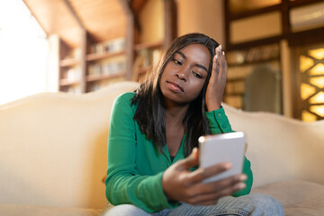 Unhappy young black lady reading depressing news on cellphone, receiving bad message at home