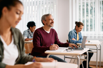 Happy mature man uses laptop and takes  notes during lecture in classroom.