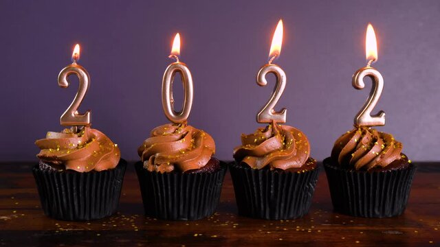 Happy New Year's Eve 2022 chocolate cupcakes decorated with gold burning candles on aqainst dark wood table setting background. Cinemagraph static loop.