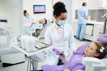 African American dentist holds little girl's hand and consoles her during dental procedure at dentist's office.