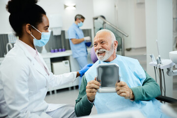 Satisfied senior man looks his teeth in a mirror and talks with his dentist at dental clinic.