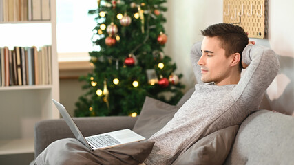 Man sitting on comfortable couch near Christmas tree and using laptop computer.