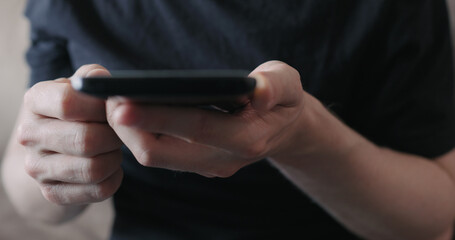 Closeup young man in black t-shirt typing messages on smartphone while sitting on a couch