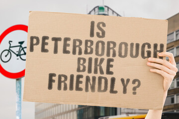 The question " Is Peterborough bike friendly? " on a banner in men's hand with blurred background. Transportation. Zero waste. Bicycle lane. Streets. City. Safety. Insecure. Road signs. Dangerous