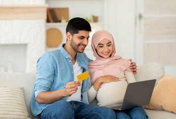 Preparing for childbirth. Loving muslim pregnant couple using laptop computer and credit card, sitting on sofa