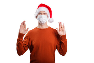 an isolated shot of a surprised man in a medical mask, in casual clothes, a Christmas hat cap. He raised his hands, crossed his fingers, looked at the camera